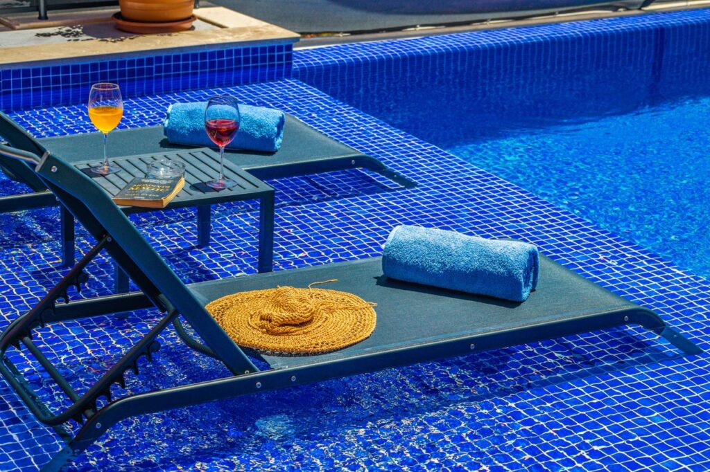 Enjoy a tranquil poolside scene with deckchairs, towels, and refreshing drinks beside a sparkling blue pool.
