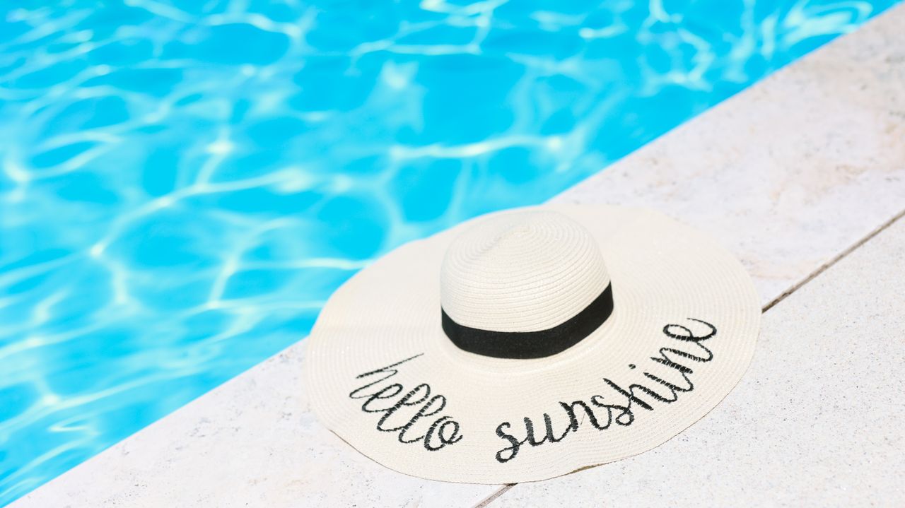 A beige straw hat with 'hello sunshine' text by a sparkling blue swimming pool.